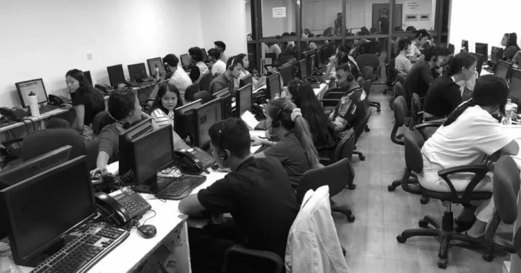 Wide black-and-white view of an RHI support floor with agents working at computers and headsets.